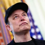 Elon Musk at Tesla shareholder meeting with Tesla vehicles and robot prototype in background.